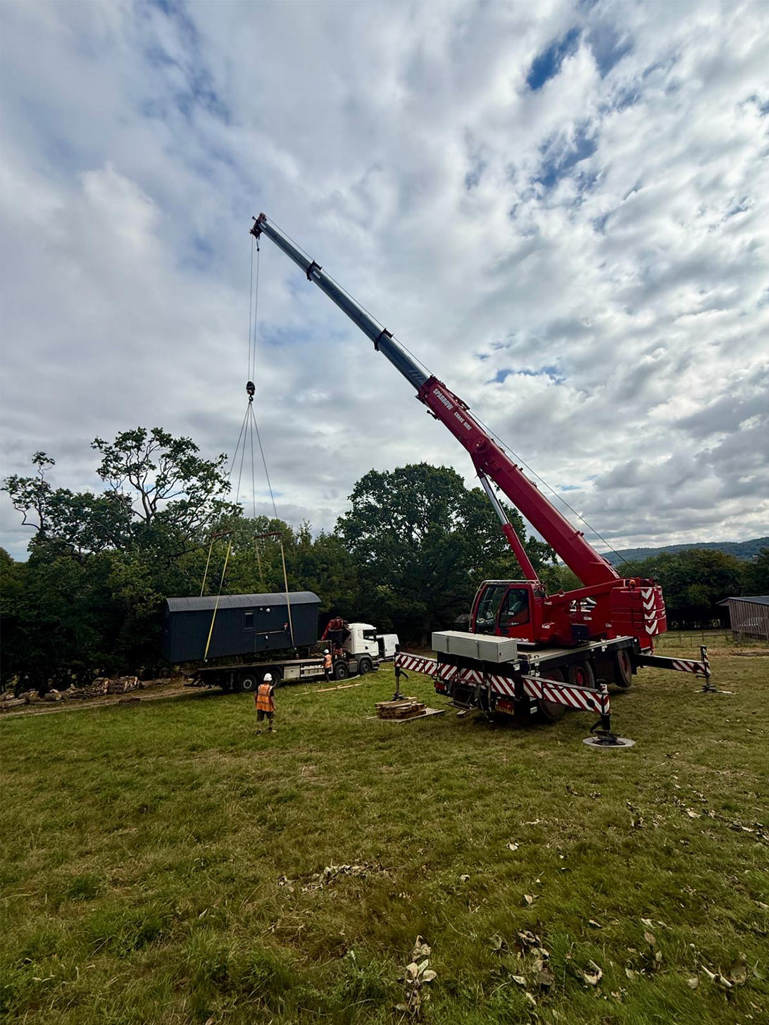 Liebherr mobile crane using outriggers to lift a shepherd hut across sloping ground during installation in the South West